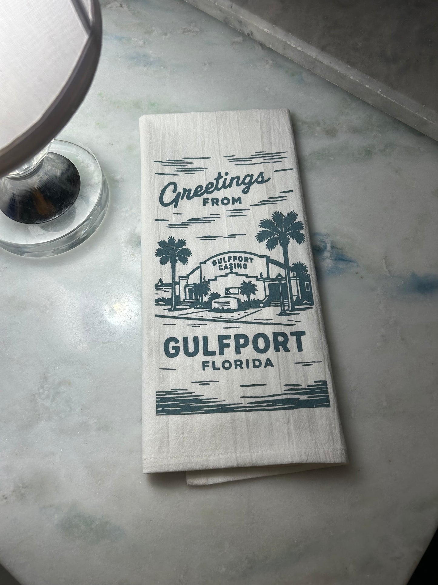 Folded white flour sack towel with 'Greetings from Gulfport, Florida' text and an image of Gulfport Casino, on a marble surface